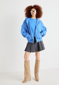 Blå zip-up hettegenser, blå crew neck t-skjorte, plissert svart miniskjørt, beige knelengde støvler, sølvsolbriller. Myk tekstur, avslappet design.
