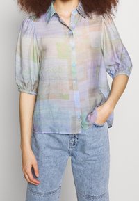 Blusa ligera de color pastel con botones en el frente, mangas cortas abullonadas y un ajuste ligeramente holgado. Presenta un patrón abstracto.