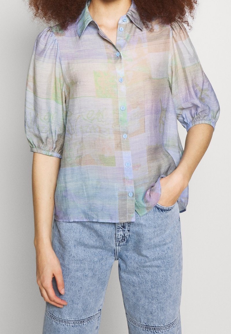 Blusa ligera de color pastel con botones en el frente, mangas cortas abullonadas y un ajuste ligeramente holgado. Presenta un patrón abstracto.