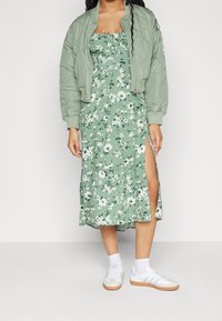 Zielona kwiatowa sukienka z rozcięciem do uda, w zestawie z jasnooliwkową kurtką bomberką i białymi sneakersami na gumowej podeszwie.