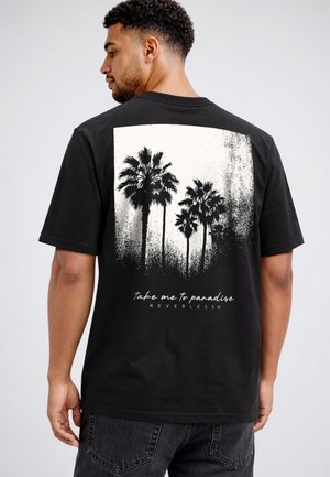Man wearing black t-shirt with black and white palm tree graphic and text "take me to paradise NEVERLESS" on the back.