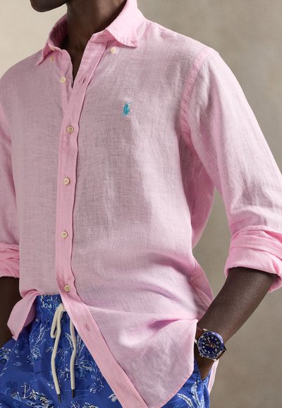 Ροζ λινό πουκάμισο με κουμπιά και μπλε λογότυπο, συνδυασμένο με μπλε σορτς με σχέδια και μπλε ρολόι. Υφασμάτινη υφή, άνετη γραμμή, με τυλιγμένα μανίκια.