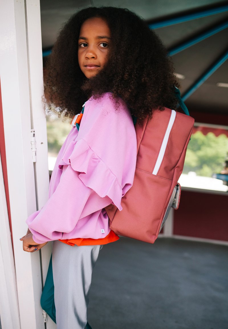 Fille aux cheveux bouclés portant une veste rose et un sac à dos rose, debout près d'une porte dans un espace extérieur ombragé.