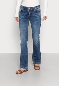 Femme portant un jean bootcut bleu, un haut blanc à manches longues côtelé et des sandales plates marron, debout sur un sol gris devant un mur blanc.