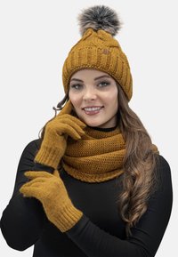 Gorro de punto amarillo mostaza con pompón de piel, bufanda a juego con patrones texturizados y guantes, todos con puños acanalados y textura suave.