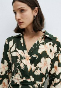 Tamnozeleni bluzon s floralnim uzorkom u breskvastoj i bijeloj boji. Ima ovratnik, duge rukave i detalj oko struka koji se vezuje.