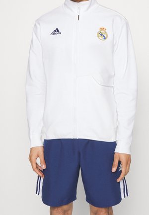 Person iført en hvid Adidas lynlåstrøje med Real Madrid-logo og mørkeblå Adidas-shorts med hvide striber.