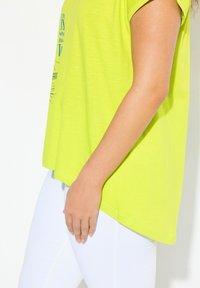 Camiseta amarilla neón brillante con un corte holgado, mangas cortas, dobladillo curvado y texto gráfico azul, combinada con pantalones blancos ajustados.