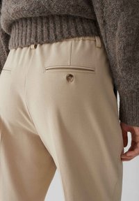Beige broek met een gladde textuur, voorzien van twee achterzakken met klep en knoopdetail, gecombineerd met een getextureerde bruine sweater.