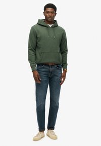 Felpa verde con cappuccio e stringhe, tasca anteriore, indossata con jeans blu e sneaker crema. Design semplice e vestibilità rilassata.