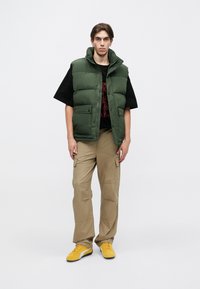 RohelinePaddingutega vest kõrge kraega, must T-särk punase graafilise kujundusega, beežid cargo-püksid ja kollased spordijalatsid.