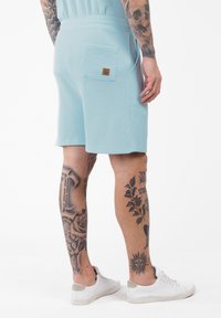 Uomo che indossa pantaloni corti azzurri chiari con texture e sneakers bianche, con ampie tatuaggi in inchiostro nero su polpacci e avambracci, in piedi su uno sfondo bianco.