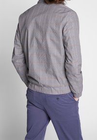 Mężczyzna ubrany w jasną, kratkowaną marynarkę i niebieskie spodnie, z rękami w kieszeniach, pokazany od tyłu na jednolitym tle.