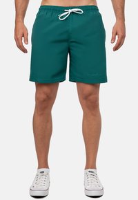 Pantaloni da bagno turchesi con vita elastica, lacci bianchi e tasche laterali. Presentano un orlo cucito e un dettaglio di branding discreto.