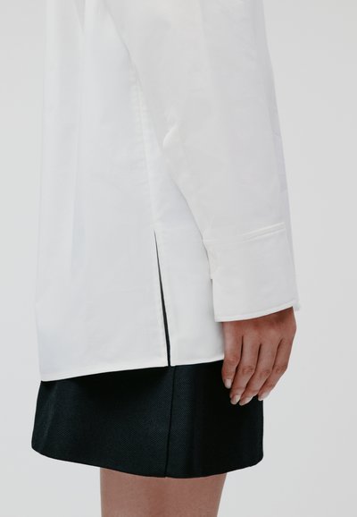 White long-sleeve shirt with a side slit, featuring buttoned cuffs. Paired with a black textured skirt. Clean, minimalist design.