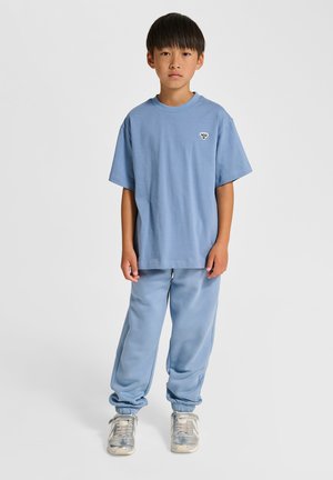 Junge steht, trägt hellblaues oversized T-Shirt und passende Jogginghose mit silbernen Sneakern, schlichter heller Hintergrund.