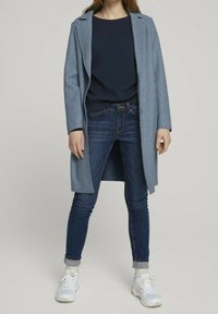 Manteau bleu clair avec revers sur un pull texturé bleu marine, associé à un jean foncé et des baskets blanches. Tenue décontractée aux lignes épurées.