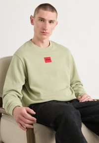 Sweatshirt vert clair avec un col rond et un logo carré rouge portant le nom "HUGO", associé à un jean foncé. Texture lisse, coupe décontractée.