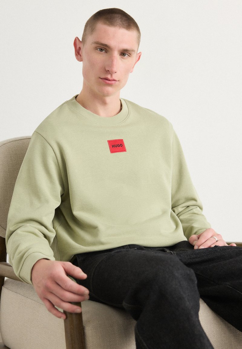 Sweatshirt vert clair avec un col rond et un logo carré rouge portant le nom "HUGO", associé à un jean foncé. Texture lisse, coupe décontractée.