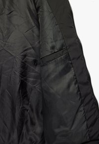 Indersiden af en sort quiltet jakke med en glat, skinnende tekstur, der har en sidelomme og en lynlås på den forreste kant.