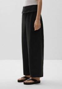 Pantalons noirs taille large avec un waistband replié, fabriqués en tissu léger. Portés avec des tongs noires, montrant un design épuré et minimaliste.