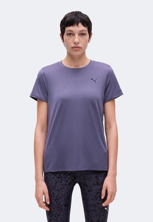 Młoda osoba z krótkimi ciemnymi włosami, ubrana w fioletowy t-shirt Puma i czarne legginsy w wzory, stoi na jasnym tle.
