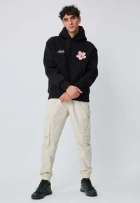 Sweat à capuche noir avec un motif de fleur rose sur la poitrine, associé à un pantalon cargo beige avec plusieurs poches et des poignets élastiques. Chaussures noires.