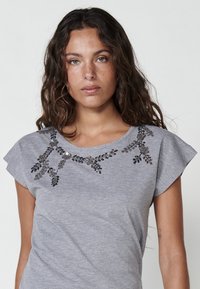 Grå kortärmad t-shirt med ett dekorativt mönster av pärlor och paljetter runt kragen, mjuk textur och avslappnad passform.