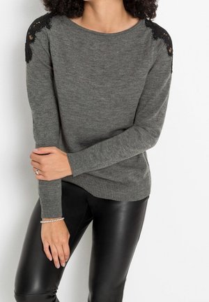 Femme portant un pull gris avec des détails en dentelle noire sur les épaules et un pantalon en cuir noir, debout sur fond blanc.
