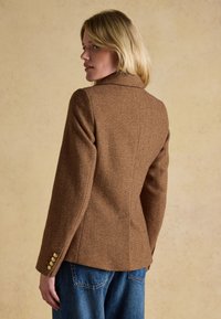 Femme blonde portant un blazer marron en chevrons taillé sur mesure avec des boutons dorés aux poignets, associé à un jean en denim bleu, debout de profil.