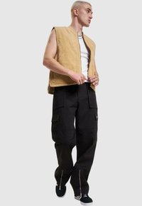 Gilet en velours côtelé beige sans manches, associé à un pantalon cargo noir avec poches et zippers, porté avec des baskets noires.