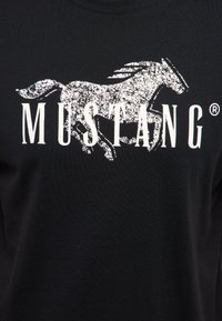 Hvidt, slidt grafisk billede af en løbende hest over ordet "MUSTANG" i fete, store bogstaver på en sort stofbaggrund.