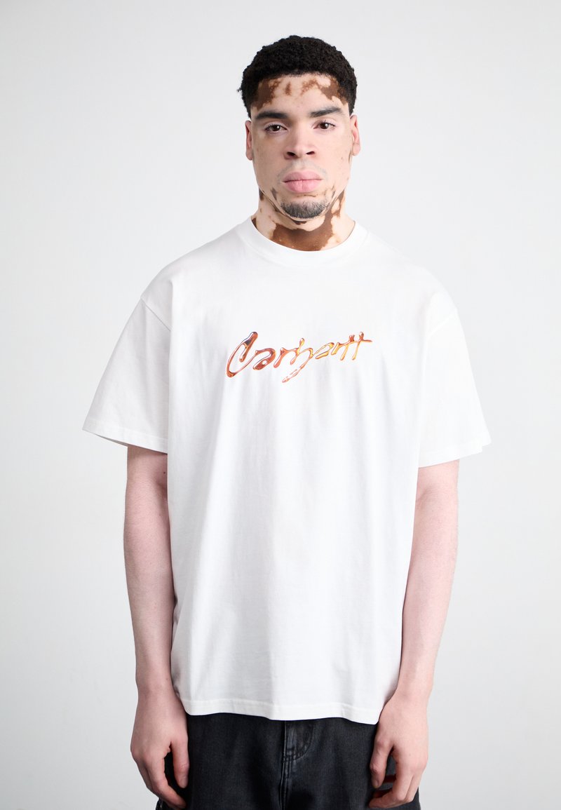 Witte T-shirt met korte mouwen met het "Carhartt" logo in oranje en bruin op de voorkant, gecombineerd met donkere broek, tentoongesteld tegen een effen achtergrond.