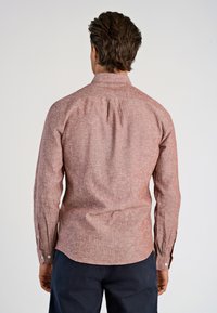 Man med mörkt hår som bär en långärmad, knäppt, röd texturerad skjorta och mörka byxor, sedd bakifrån mot en vit bakgrund.