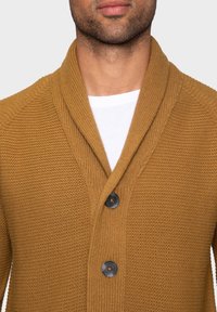 Musztardowy sweter na drutach z kołnierzem szalowym, teksturowaną tkaniną i trzema czarnymi guzikami do zapięcia.