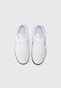 Sapatilhas Nike brancas com um superior de couro liso, bico redondo, gola acolchoada, detalhes em prata e atacadores brancos sobre uma língua cinza. Sola de borracha preta.