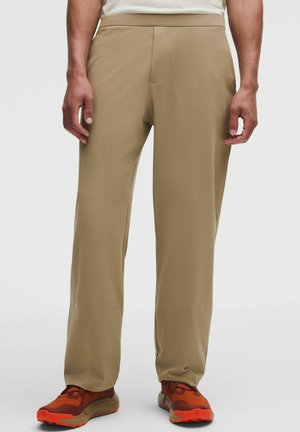 Mand iført beige bukser med lige ben og orangebrune sneakers, stående med hænderne afslappet ned langs siderne på hvid baggrund.