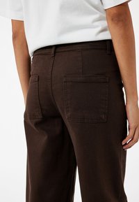 Pantalones de mezclilla marrones con un ajuste de cintura alta, que cuentan con dos bolsillos traseros y un acabado limpio. La textura es suave con costuras mínimas visibles.