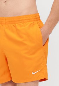 Narancssárga úszónadrág, rugalmas derékkal, oldalsó zsebbel és fehér Nike logóval a bal alsó oldalon. Simító, könnyű anyag.