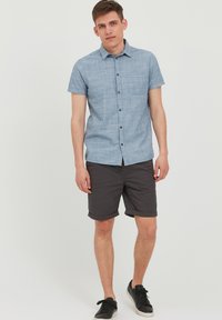 Kortærmet, lyseblå skjorte med knapper og et struktureret mønster samt en brystlomme, parret med rullede mørke shorts og sorte sneakers.