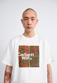 Biela bavlnená tričko s štvorcovou grafikou červených kvetov na zelenom pozadí, s nápisom "Carhartt WIP" v tučnom bielom písmene uprostred.