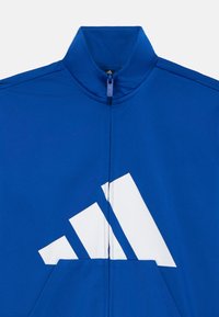 Veste zippée bleue avec un col haut et un grand logo Adidas blanc avec trois rayures diagonales. Tissu lisse et texturé.