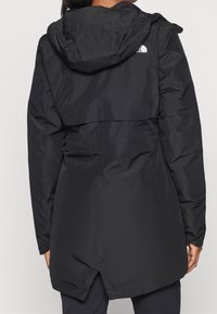 Manteau noir imperméable avec capuche, présentant une taille cintrée, de longues manches et un ourlet fendu. Le tissu a une finition lisse et brillante.