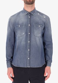 Camicia in denim di colore blu chiaro con due tasche sul petto, leggermente usurata e maniche lunghe. Presenta un colletto classico e bottoni frontali.