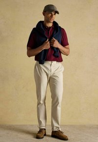 Burgundi polo särg, kreemjad püksid, tumesinine kardigan õlgadel, pruunid lipsudega kingad, houndstooth müts, seismine neutraalse tausta ees.