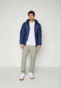Jachetă matlasată bleumarin cu glugă și logo pe piept, tricou alb, pantaloni cargo verzi deschis și adidași albi cu talpă maro.