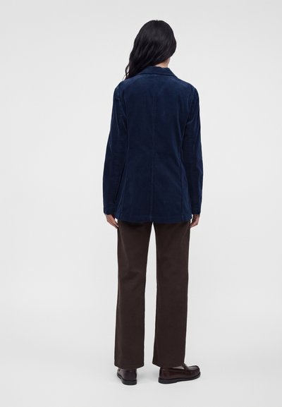 Femme debout, vue de dos, portant une veste bleu foncé, un pantalon droit marron et des chaussures marron foncé sur un fond uni.