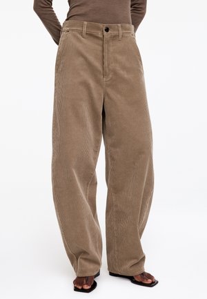 Persona que lleva pantalones de pana beige de talle alto y pierna ancha con bolsillos delanteros y sandalias negras abiertas en los dedos, sobre un fondo liso.