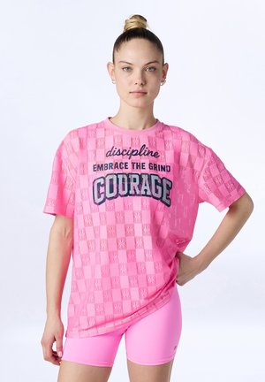 Giovane donna con capelli raccolti in un bun, indossa una maglietta rosa acceso con la scritta "disciplina abbraccia la fatica coraggio" e pantaloncini rosa coordinati, in piedi con una mano sul fianco.