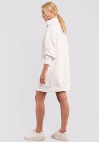 Abito felpa bianco oversize con colletto alto, polsi a coste e tessuto liscio, indossato sopra sneakers bianche.